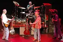Photographie d'un groupe de rock composé de quatre personnes. L'un est au fond devant une batterie, les trois autres sont au premier plan, un à gauche avec une guitare, un au milieu avec une guitare et devant un micro et le troisième au fond avec une basse.