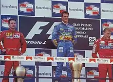 Photo de trois hommes, deux en rouge, aux côtés d'un homme en bleu, sur un podium, et devant un mur de sponsors