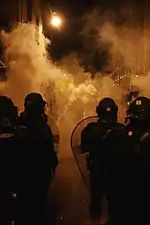 Plusieurs policiers en uniforme, casqués, tenant boucliers. Vêtements sombres et feu en arrière-plan.