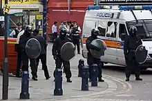 Quatre policiers en uniforme noir et casque, tenant boucliers ronds transparents sur un coin de rue. Plusieurs véhicules et piétons en arrière-plan.