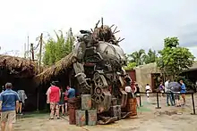 Une machine rouillée constituée de deux bras et de deux jambes devant une cabane en bois.