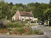 Au premier plan : vue d'un rond-point et de son terre-plein fleuri. Au second plan : vue d'un maison. À l'arrière-plan : les premiers arbres du bois de Sarron.