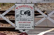 Gros plan sur le panneau émaillé du portillon qui porte l'indication : « NE TRAVERSEZ PAS sans regarder dans les DEUX DIRECTIONS. Un train peut en CACHER un autre » et un dessin avec une famille qui traverse alors que survient une locomotive à vapeur qui leur était masquée par un wagon.