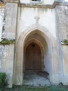 photo couleur d'un porche en arc brisé surmonté d'une petite croix sculptée dans une pierre du mur. Il est encadré de deux contrefort. Le porche est dallé de pierres plates et la porte au fond est en bois à deux battants et un oculus.