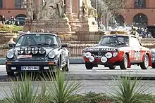 Photo d'une Porsche 911 et d'une Lancia Fulvia, roulant en ville et longeant une fontaine.