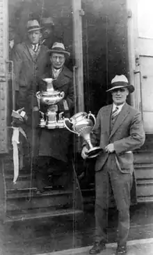 Photographie en noir et blanc de trois hommes descendant d'un train, deux d'entre eux tenant une coupe dans la main