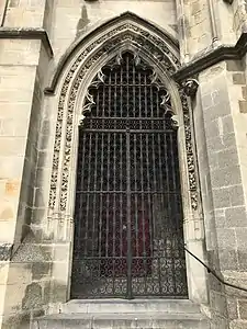 Porte fermée par une grille en fer forgé et entourée de volutes sculptées.