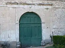 photo couleur montrant une porte en bois à deux battants peinte en vert délavé. Le linteau en arc de cercle est surmonté par une corniche supportée par les pilastres qui encadrent la porte.