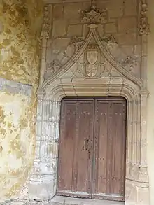 Photo couleur d'une porte en bois cloutée à deux ventaux. Les montants en pierre sont nervurés et le linteau est surmonté d'un arc brisé orné de blasons.