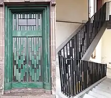 Photographie en couleurs représentant à gauche une porte d’entrée en ferronnerie verte et à droite un escalier blanc avec une main courante en ferronnerie noire.