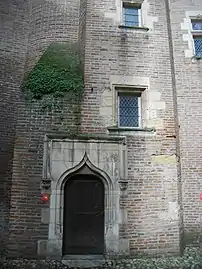 Photo couleur d'une porte à encadrement de pierre ouvragé dans une bâtisse de briques rouges. Le linteau est en anse de panier à décoration d'arc brisé très pointu. Une fenêtre à petits carreaux en losange est située au-dessus de la porte à droite.