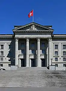 Photographie d'un monument, au premier plan un grand escalier, au centre quatre grandes colonnes encadrant les portes d'entrée, le toit en v comprend une statue en son centre et est surmonté d'un drapeau rouge à croix blanche