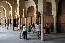 Photographie d'un groupe de touristes dans le portique à double travée, précédant la salle de prière. Celle-ci est entièrement fermée, côté portique, par des portes en bois sculpté.