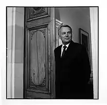 Photo en noir et blanc de Jean-Marc Ayrault, en costume noir, se tenant dans l'embrasure d'une porte.
