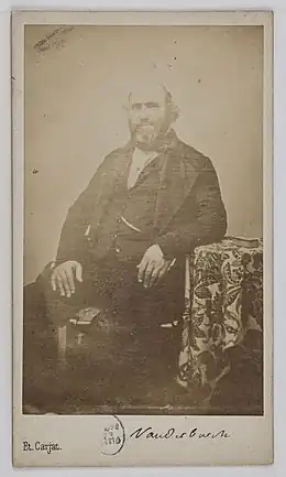 Photographie en noir et blanc représentant un homme mûr barbu en costume trois pièces sombre assis de trois-quart face sur un fauteuil et accoudé à un guéridon recouvert d'un tissu.