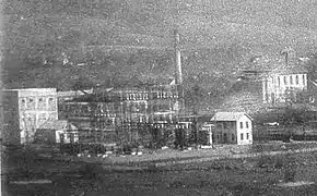 Photo noir et blanc montrant des bâtiments trapus et des structures métalliques, derrière se trouve une grande cheminée et des bâtiments de charbonnage