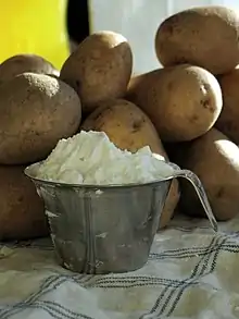 Pichet métallique rempli d'une farine blanche, posé sur un torchon blanc à carreaux bleus, devant un pile d'une dizaine de pommes de terre entières