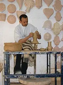 Potier de Guellala travaillant de l'argile sur son tour ; le mur derrière lui est couvert de poteries.