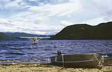 Photographie couleur d'un petit bateau sur une plage et d'un hydravion amerissant sur l'eau.