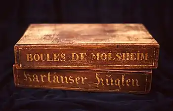 Deux boîtes de bois superposées, sur lesquelles sont écrits respectivement « Boules de Molsheim » et « Kartäuser Küglen.