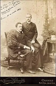 Photo en noir et blanc de deux hommes en habit et redingote, dont l'un est assis sur un fauteuil sur l'accoudoir duquel le second est assis.