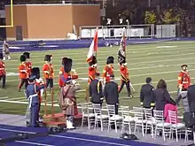 Photographie en couleur de militaires en uniformes écarlates tenant des drapeaux et défilant devant des dignitaires les saluant