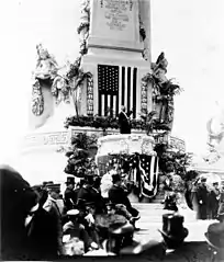 Photographie en noir et blanc d'un homme prononçant un discours devant un drapeau américain