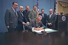 Un homme, assis, signant un document, six hommes, debout, derrière lui.