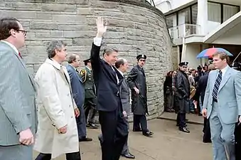 Un homme en costume lève la main en signe de salut. Il est entouré par plusieurs hommes en costume.