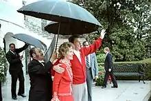 Un homme habillé d'un gilet rouge salue de la main gauche alors qu'il tient une femme, également vêtue de rouge, avec son autre main. Ils sont encadré par des agents en costume portant des parapluies.