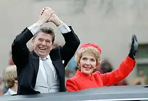 À travers le toit d'un véhicule noir, un homme en costume souriant se tient les mains au-dessus de la tête dans un signe de victoire. À ses côtés, une femme habillée de rouge salue de la main gauche.