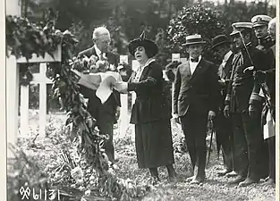 Photographie en noir et blanc d'un homme, accompagné d'une femme qui lui tend un bouquet de fleurs, et une rangée d'hommes.