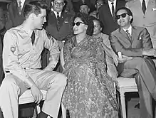 Photo en noir et blanc de trois personnes assises côte à côte : un jeune homme en uniforme militaire, en pleine conversation avec une femme en robe, et un homme en costume-cravate les bras croisés ; les deux derniers portent des lunettes de soleil