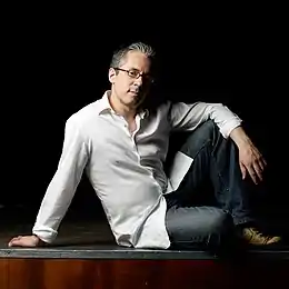 Le pianiste Manuel Rocheman, vêtu d'une chemise blanche et d'un jean, est assis près du bord de scène. Il se repose sur son bras droit, son autre bras entourant sa jambe gauche relevée. L'arrière plan est entièrement sombre.