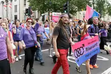 Des militants bisexuels à la marche des fiertés londonienne de 2016. Ils sont habillés couleur lavande, certains portent le drapeau bisexuel et d'autres une banderole aux couleurs bi sur laquelle il est écrit « Out and proud. Bisexuals! Fighting for equality.