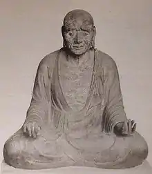 Vue de face d'une statue assise les jambes croisées avec de longs lobes d'oreilles. Ses mains reposent sur les genoux avec les paumes retournées.