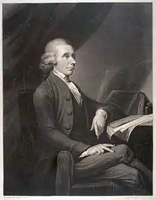 Portrait d’un homme assis sur une chaise et accoudé à une table couverte de livres et publications.