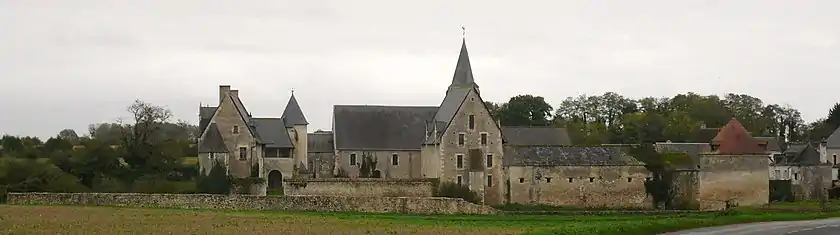 Photographie panoramique et en couleurs de bâtiments en pierre et aux toits pentus recouverts d'ardoises, un colombier à la toiture faite de tuiles rouge s'élevant sur la droite d'un pré visibles au premier plan.