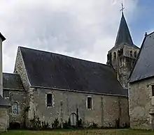 Photographie en couleurs et de profil d'une église à toit pentu, son clocher à base carrée s'élevant sur la droite, son abside s'étendant à gauche, un second bâtiment visible au premier plan.