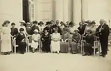 Photographie en noir et blanc. Portrait de groupe d'une vingtaine de femmes accompagnées de quelques hommes : certaines sont assises sur des chaises disposées en rang devant devant une rangée de femmes debout.