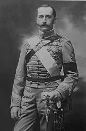 Photo de Charles de trois-quarts, en pied, l'air impassible, portant des moustaches et arborant un uniforme à brandebourgs en tenant un sabre de la main gauche