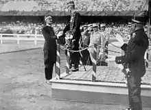 Au milieu du stade, un homme en uniforme sur une estrade serre la main d'un autre homme en uniforme; autour d'eux d'autre militaires applaudissent tandis que d'autres font le salut militaire.