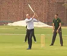 Un homme en costume tient une batte de cricket dans ses mains. Derrière lui, deux hommes se tiennent debout.