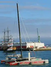 Le trimaran Prince de Bretagne vu dans l'entrée du port du Havre avec en fond un vieux gréement