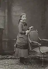 Photographie d'une fillette, à l'air pensif, en pied et de profil droit, les mains posées sur le dossier d'un fauteuil richement orné.