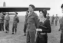 Photo en noir et blanc. Des hommes en uniforme et une femme en robe noire observent le ciel, sur un terrain herbeux.