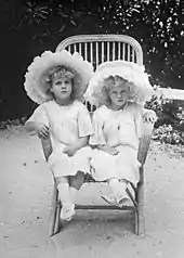 Photographie en noir et blanc de deux petites filles portant de grands chapeaux blancs et assises dans une même chaise.