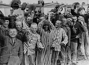 Photographie en noir et blanc représentant un groupe d'une vingtaine de prisonniers dans leur uniforme rayé. Ils se tiennent de l'autre côté d'un quadrillage de barbelés et, tout sourire, ils agitent leur chapeau à la vue des troupes américaines