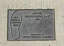 Photographie d'une plaque en metal incrustée dans le sol. Elle représente une trace de pas dans laquelle il est écrit « Bill Murray a marché ici ». Elle porte également les inscriptions en anglais « Un jour sans fin 1993, offert par Chuck Peterson & Family ».