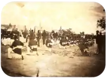 Une vieille photo montrant une procession passant entre des rangées de soldats avec des tentes en toile de fond